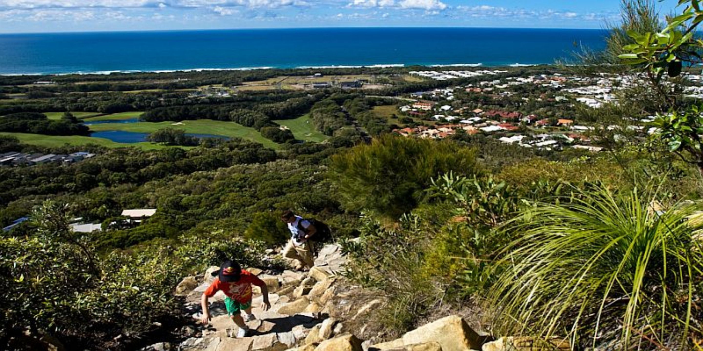 Un paisaje monta ñoso con excursionistas, vistas a un pueblo costero y una masa de agua.