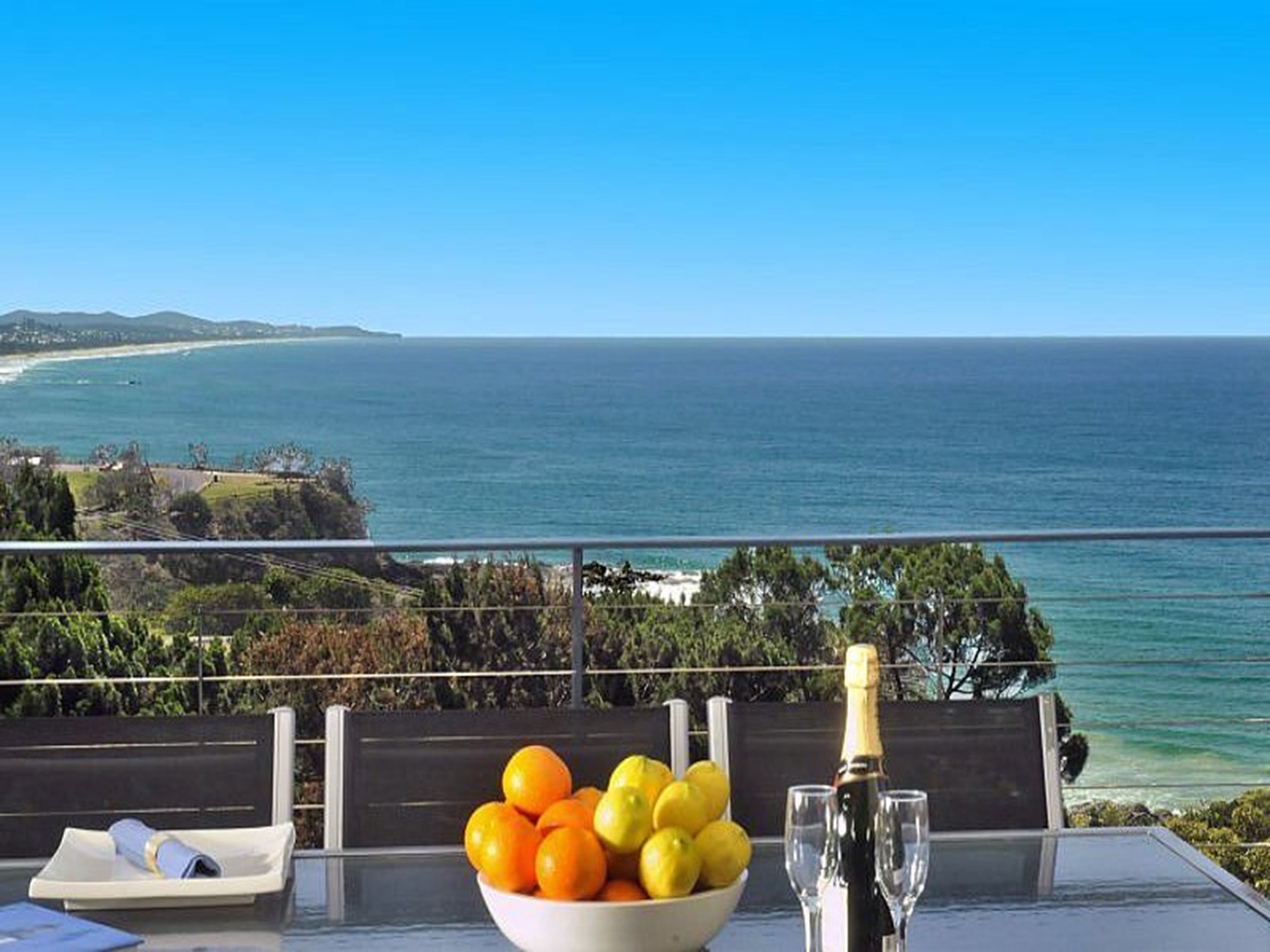 A table with a bowl of fruit, a bottle of champagne, and glasses, overlooking a coastal view.