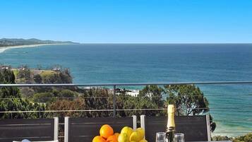 A table with a bowl of fruit, a bottle of champagne, and glasses, overlooking a coastal view.