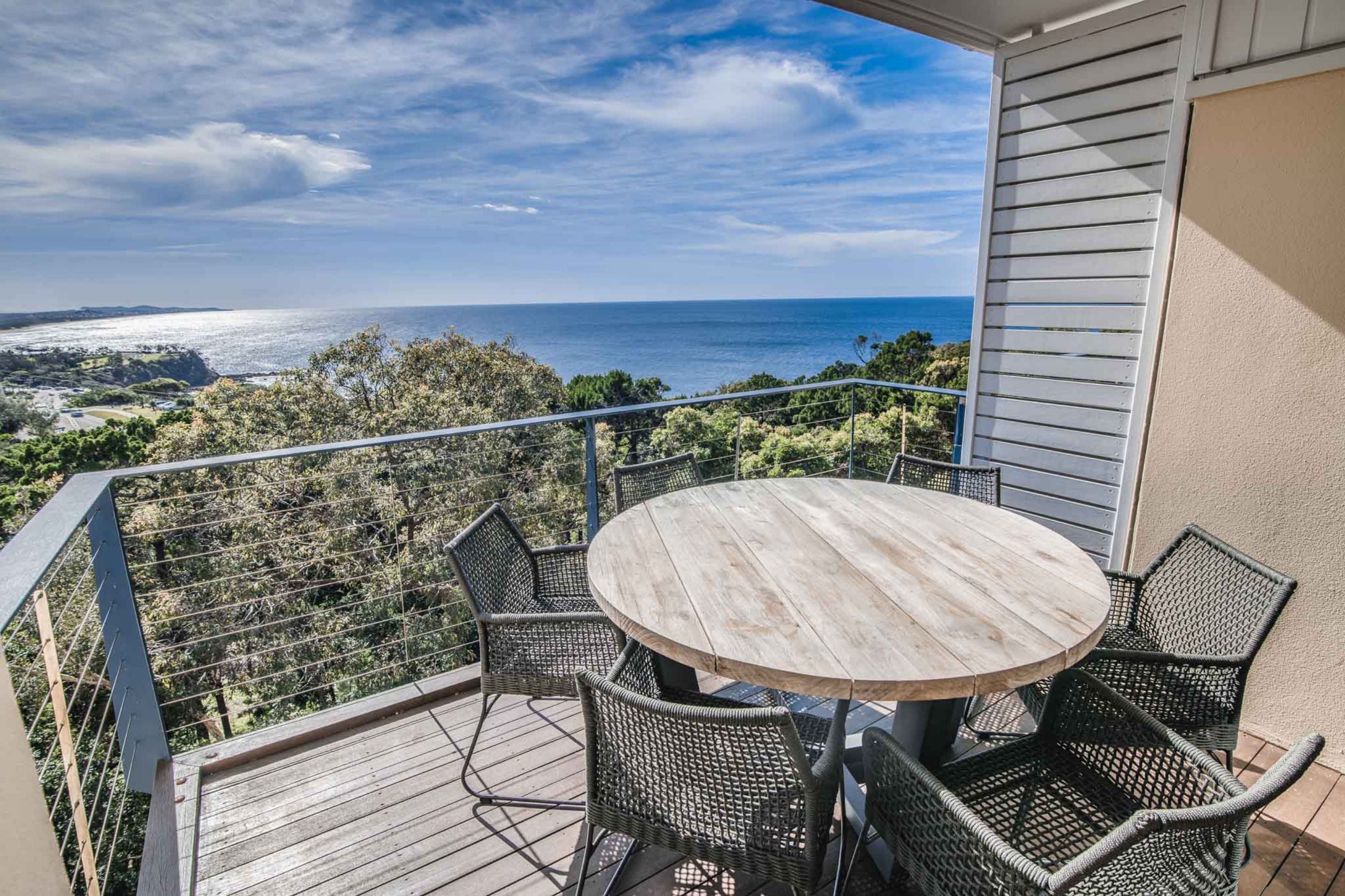 A balcony with a round wooden table and wicker chairs, overlooking a coastal landscape with a view of the sea and distant hills.