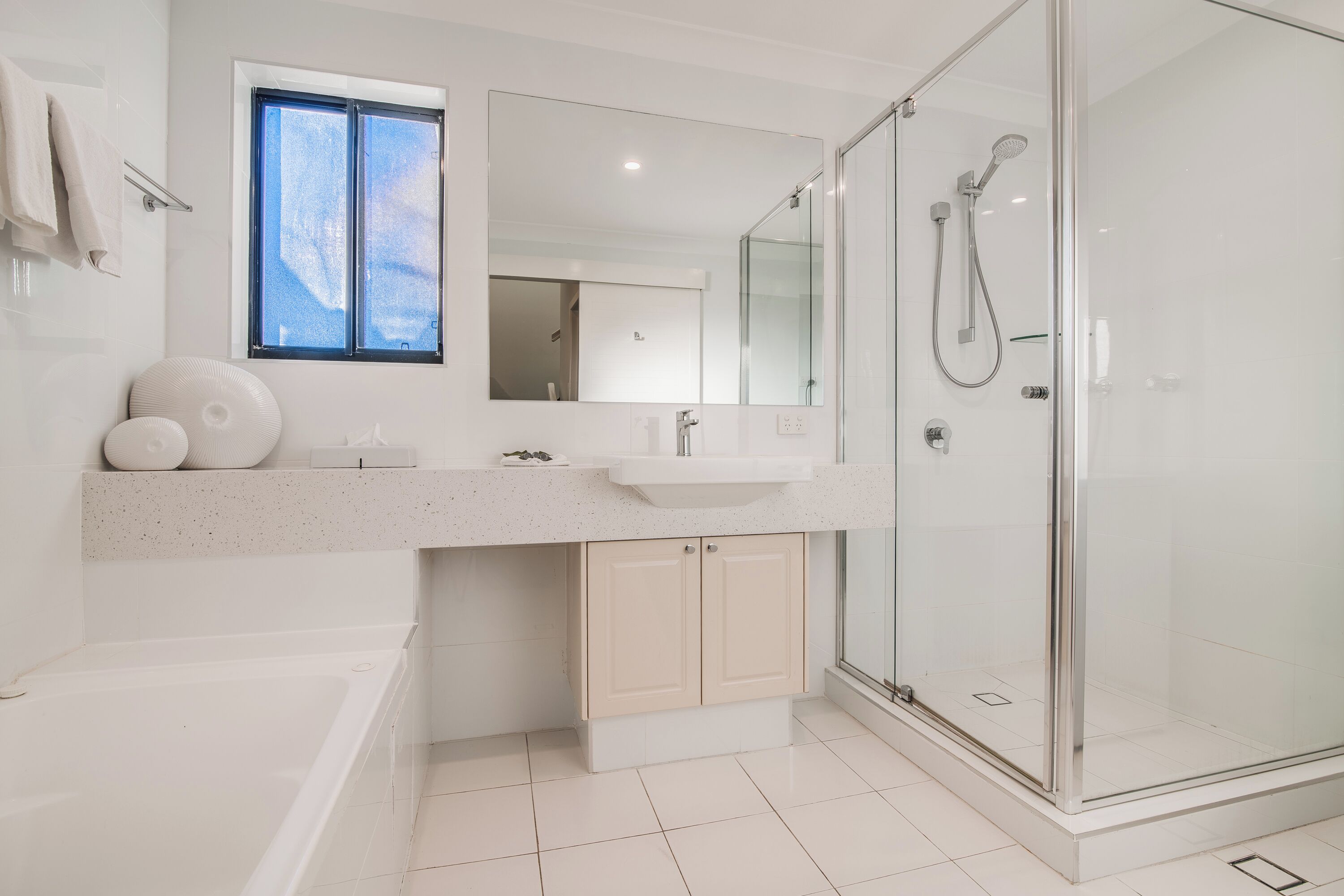 A modern bathroom with a glass shower enclosure, a bathtub, a sink with a countertop, and a window above the mirror.