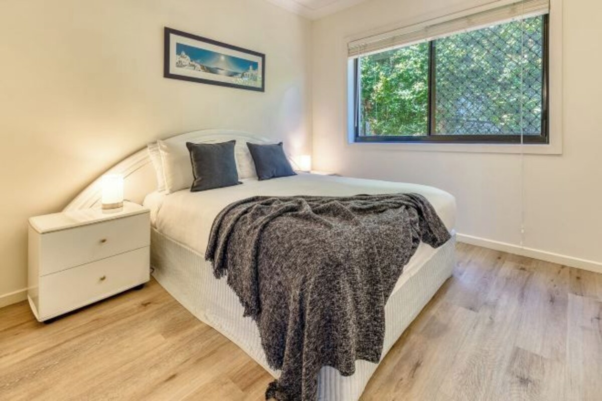 Un dormitorio con cama, mesita de noche, ventana y un cuadro en la pared.