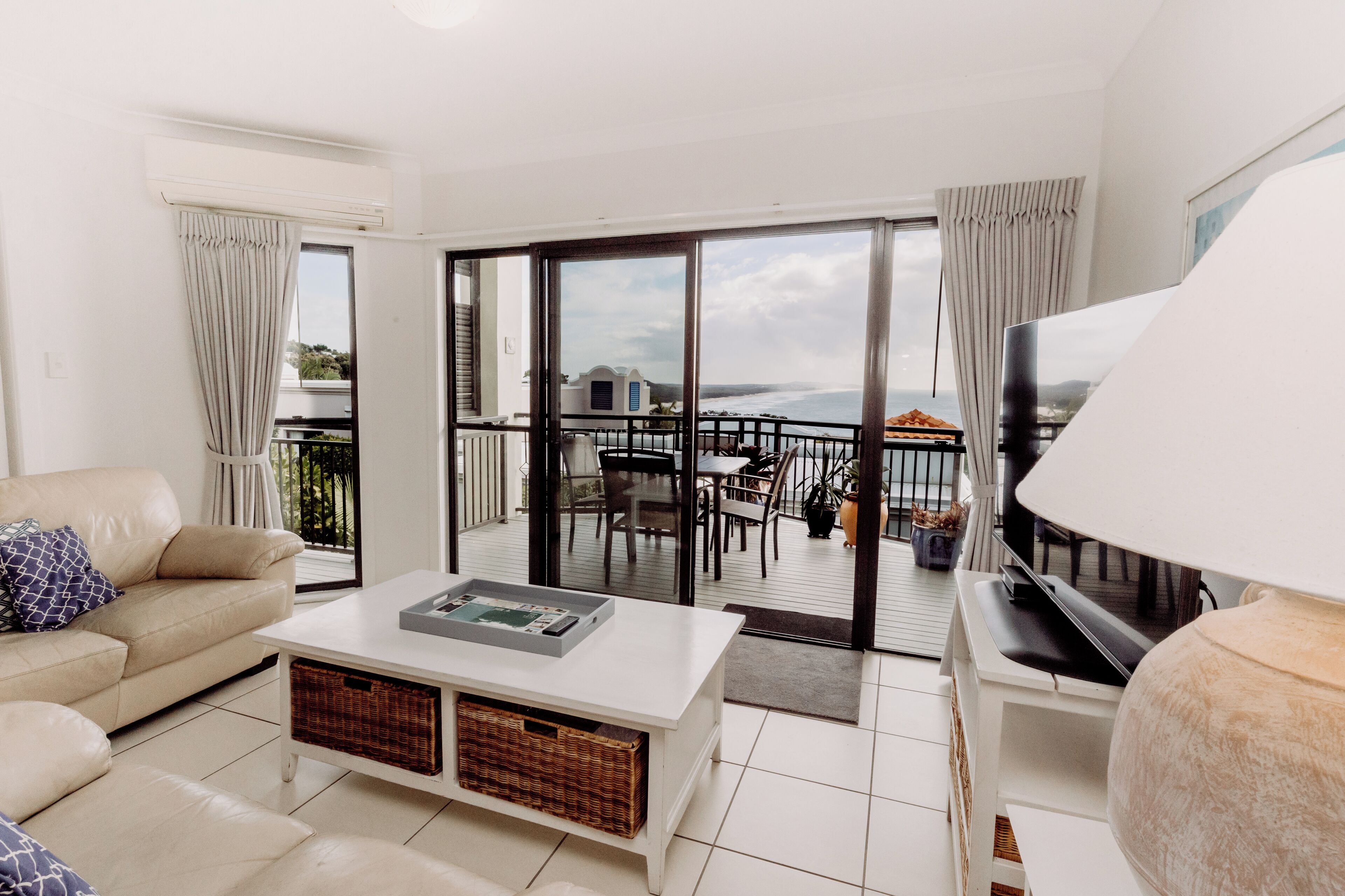 A modern living room with a sofa, coffee table, and TV. It features a balcony with a view of the sea and a dining area with outdoor furniture.