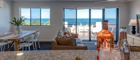 A kitchen with a granite countertop, a dining area with a table and chairs, a sofa, and a view of the ocean through large windows.