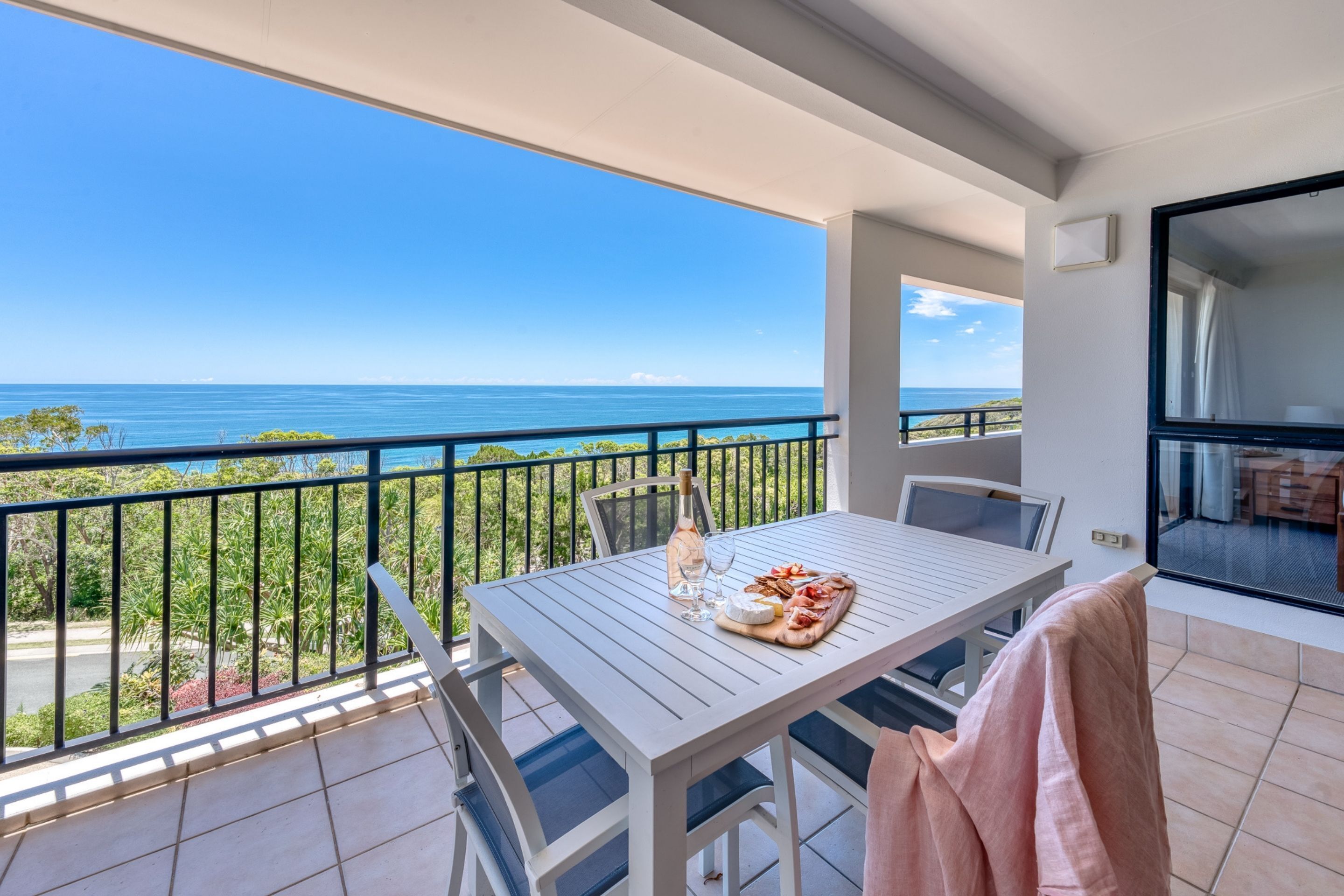 Un balcón con una mesa lista para comer, con vistas al mar azul y despejado.