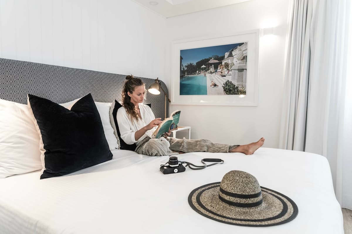 Une personne est assise sur un lit, un livre à la main, avec un appareil photo et un chapeau posés à proximité.