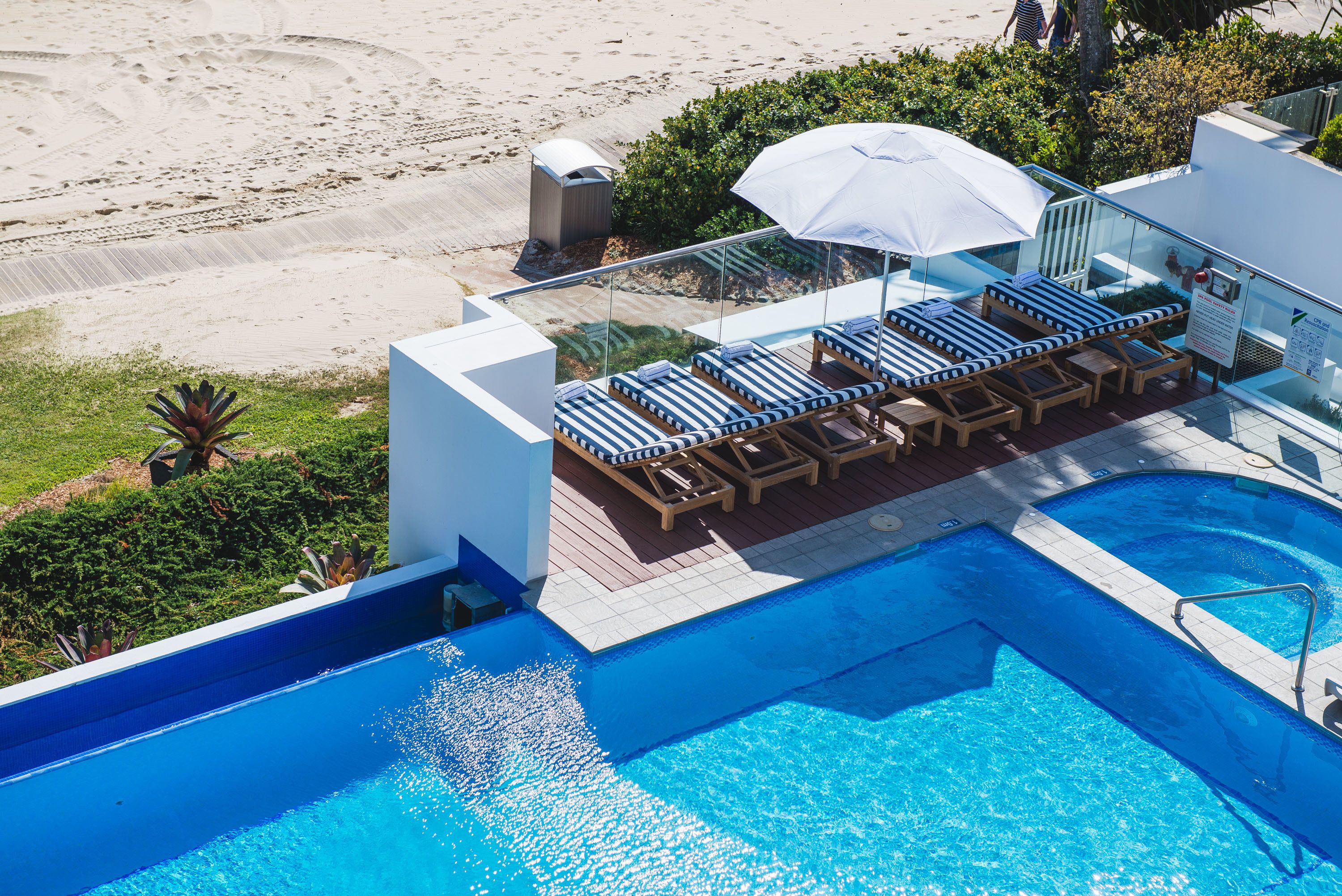 Une piscine sur le toit, dotée de chaises longues et d’un parasol, offrant une vue sur une plage de sable.