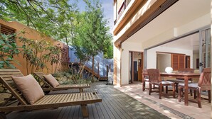 Eine Holzterrasse mit Liegestühlen und einem Esstisch, umgeben von einem Garten und einem Gebäude mit einer Schiebeglastür.