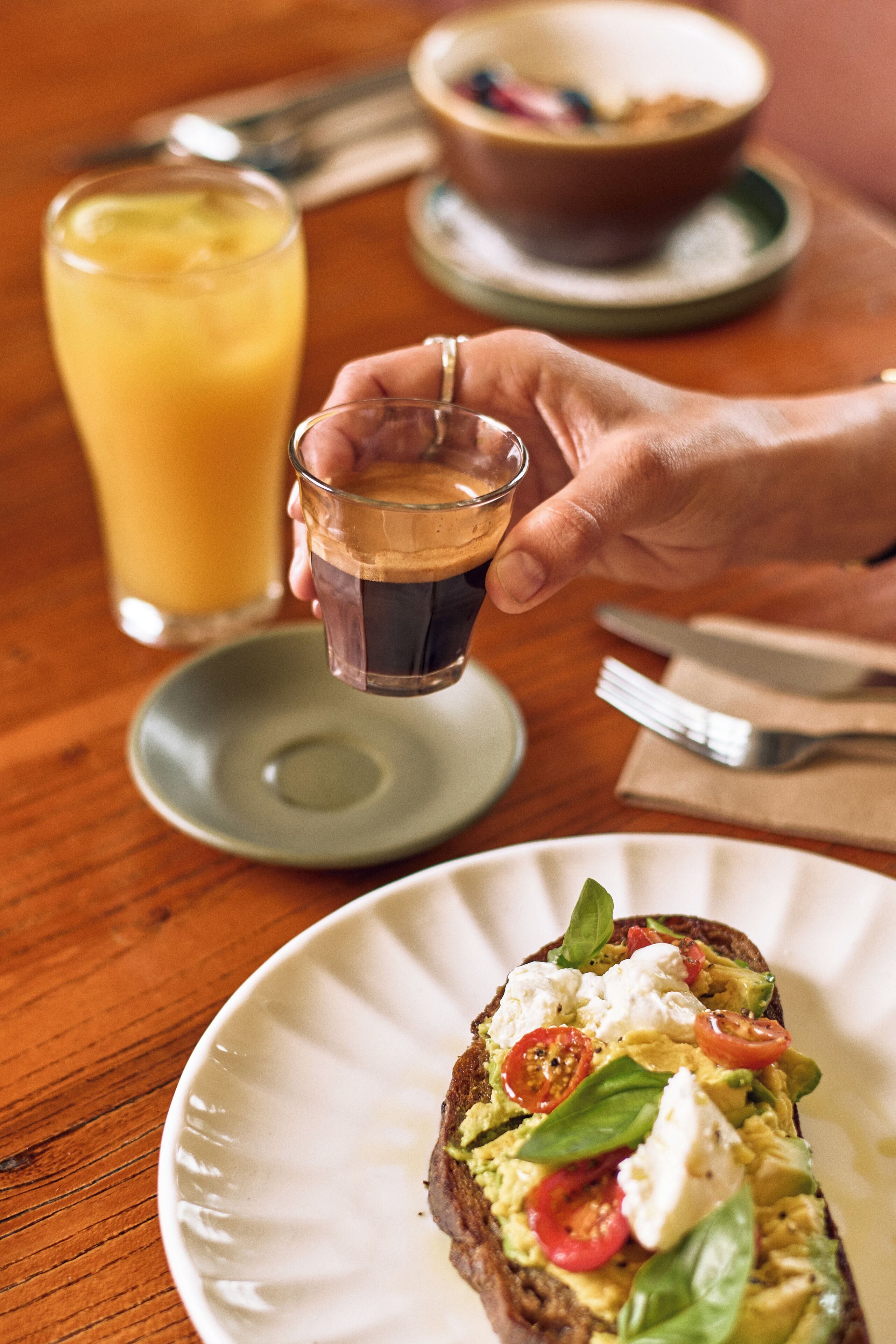Una persona sosteniendo un vaso pequeño de líquido oscuro, posiblemente café, junto a un plato con un sándwich cubierto de aguacate, tomates y hojas verdes.
