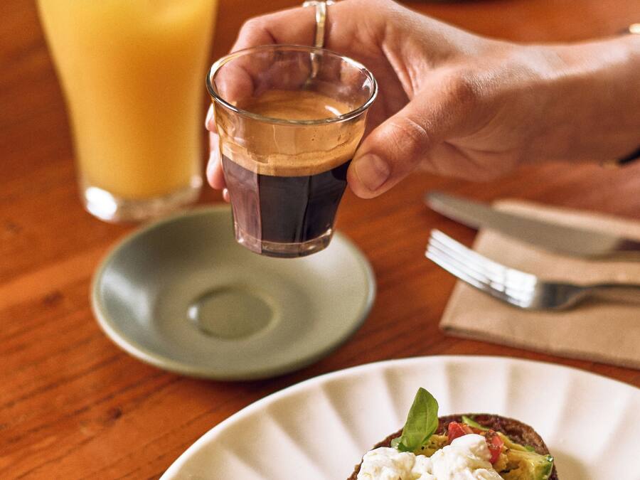 Una persona sosteniendo un vaso pequeño de líquido oscuro, posiblemente café, junto a un plato con un sándwich cubierto de aguacate, tomates y hojas verdes.