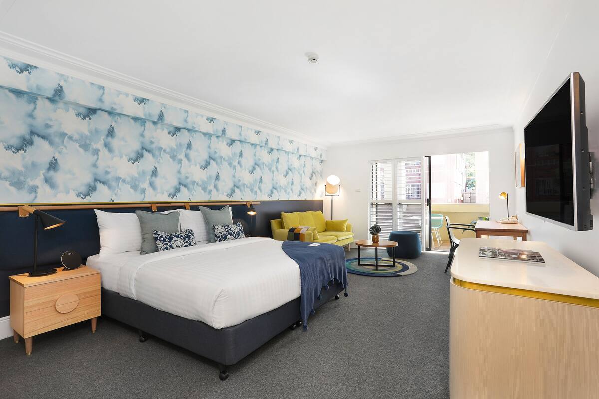 A hotel room with a bed, bedside table, lamp, and a desk with a chair.