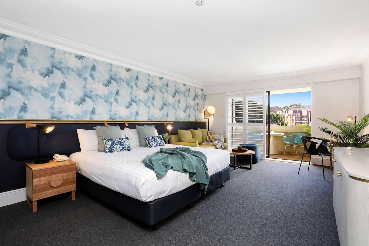 A modern bedroom with a large bed, a nightstand, a desk, and a balcony with a view.