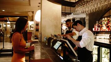 Uma área de bar com um bartender preparando bebidas, uma mulher em pé no balcão e uma adega ao fundo.