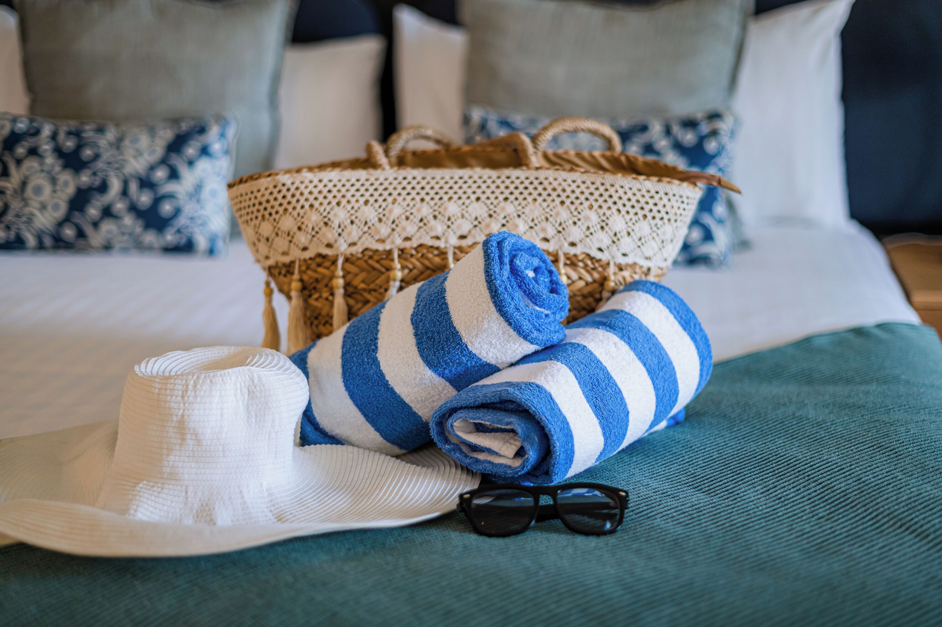 A neatly made bed with a blue and white striped towel, a white hat, and a pair of sunglasses on a teal bedspread.