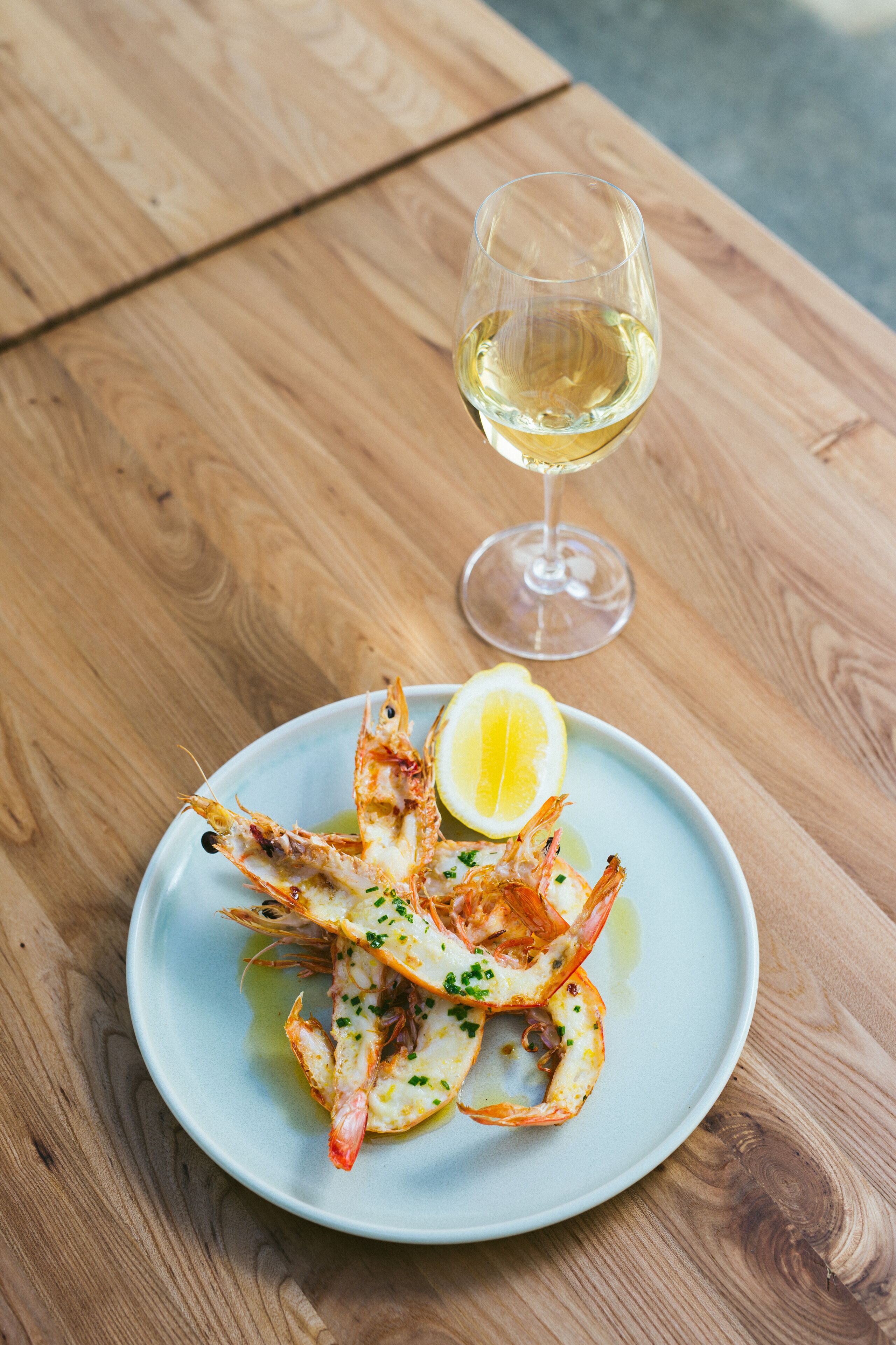A plate of cooked shrimp with lemon and herbs, accompanied by a glass of white wine on a wooden table.