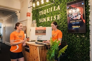Un comptoir d’accueil avec une pancarte « Bienvenue ! », une autre pancarte indiquant « Réception » et une affiche faisant la promotion de « Soirée cinéma Tequila Sunrise, chaque semaine ».