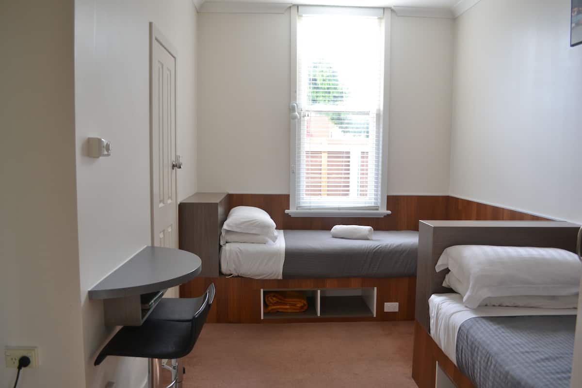Ein kleines Zimmer mit zwei Betten, einem Schreibtisch und einem Fenster mit Jalousien.