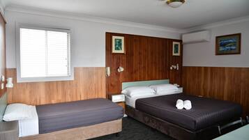 A hotel room with two single beds, a wooden headboard, a window with blinds, a bedside table, a nightstand, and a wall-mounted air conditioner.