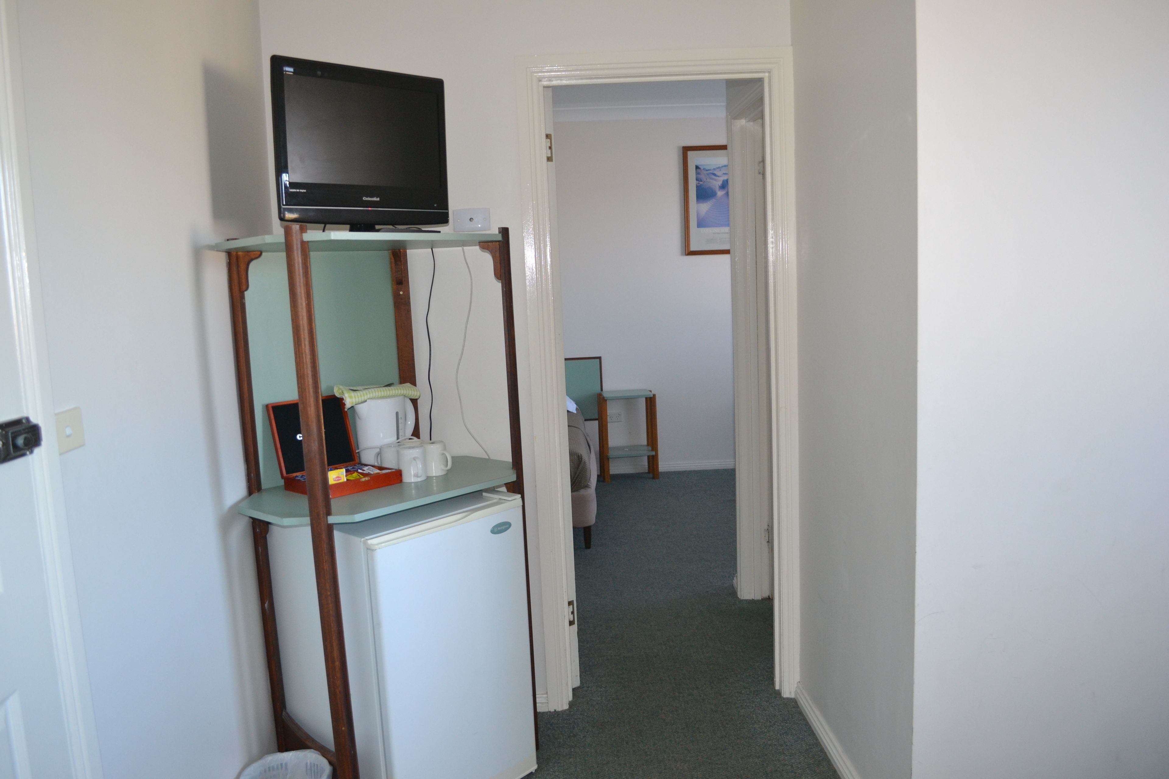 A narrow hallway with a television on a stand, a small refrigerator, and a desk with a chair.