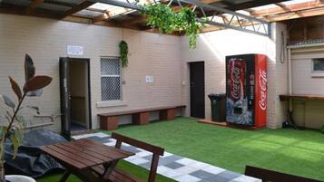 A patio area with a wooden table and benches, a Coca-Cola vending machine, and a small garden with artificial grass and potted plants.