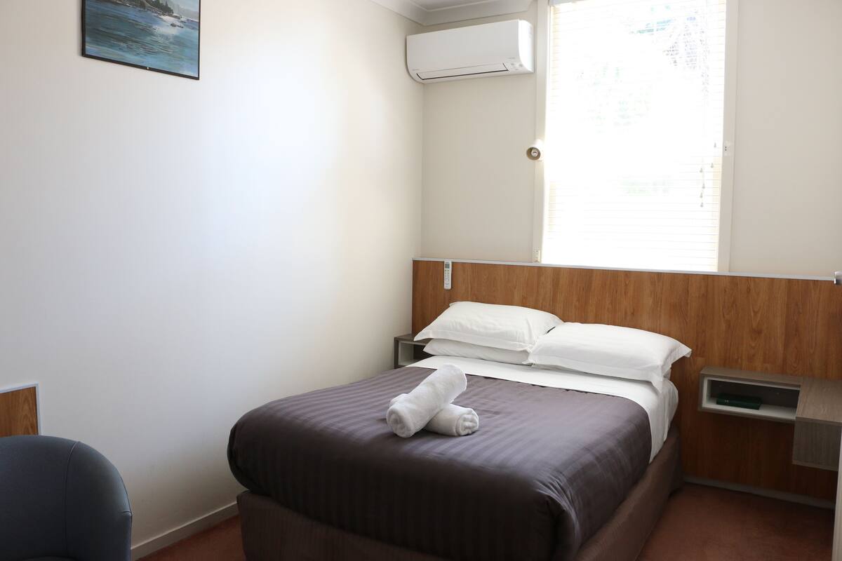 Ein ordentlich bezogenes Bett mit brauner Tagesdecke und weißen Kissen, ein Nachttisch mit Lampe sowie ein Fenster mit Jalousien.