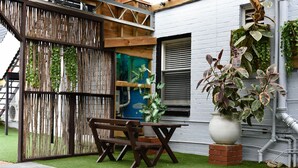 A patio with a wooden bench, a potted plant, and a white wall with a window and blinds. - Hobart Tower Motel (New Town)