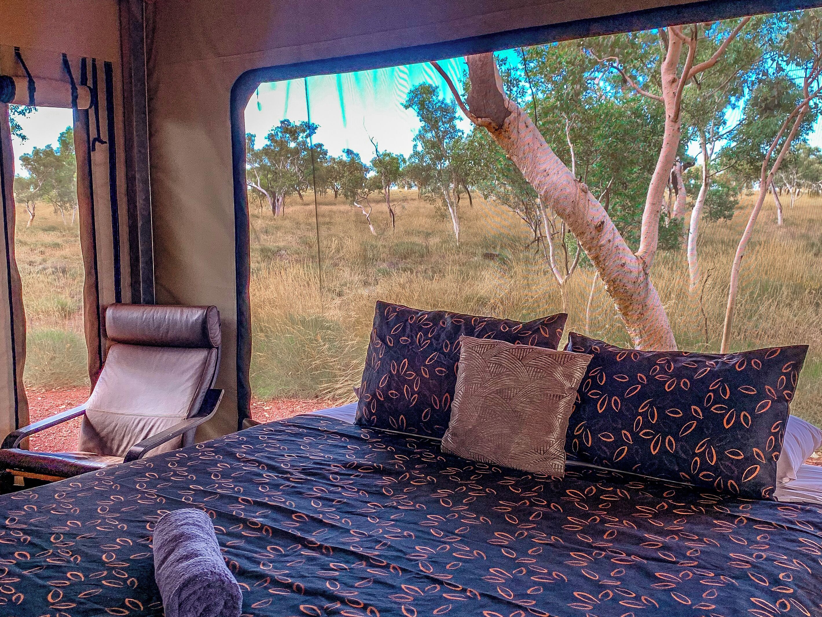 A bed with patterned bedding and pillows, a chair, and a window with a view of trees and grass.