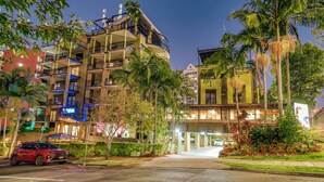 Een meerhoog hotel met balkons, palmbomen en een rode auto die ervoor geparkeerd staat.