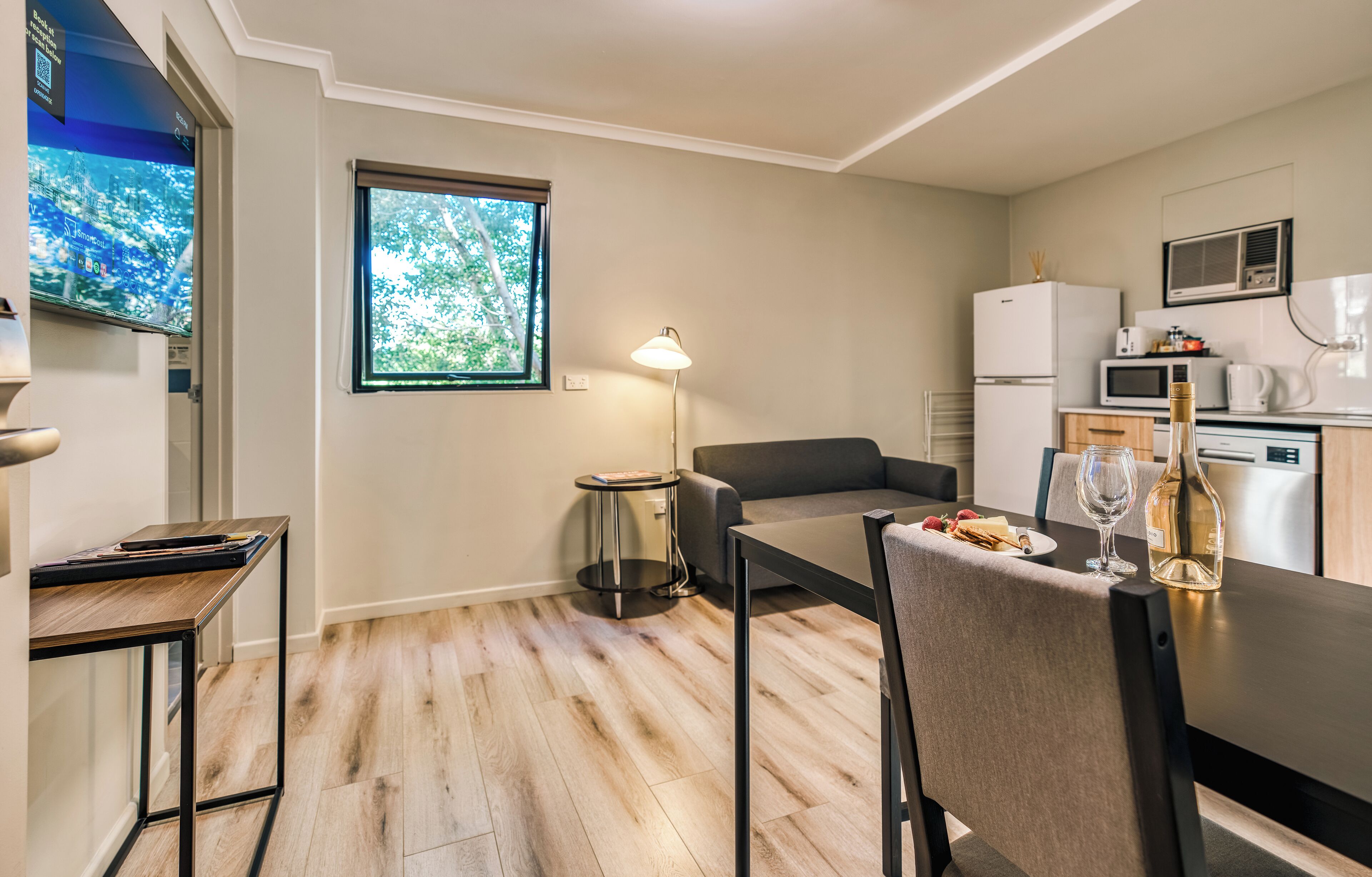 A modern kitchen with a dining area, a flat-screen TV, and a window with a view of trees.