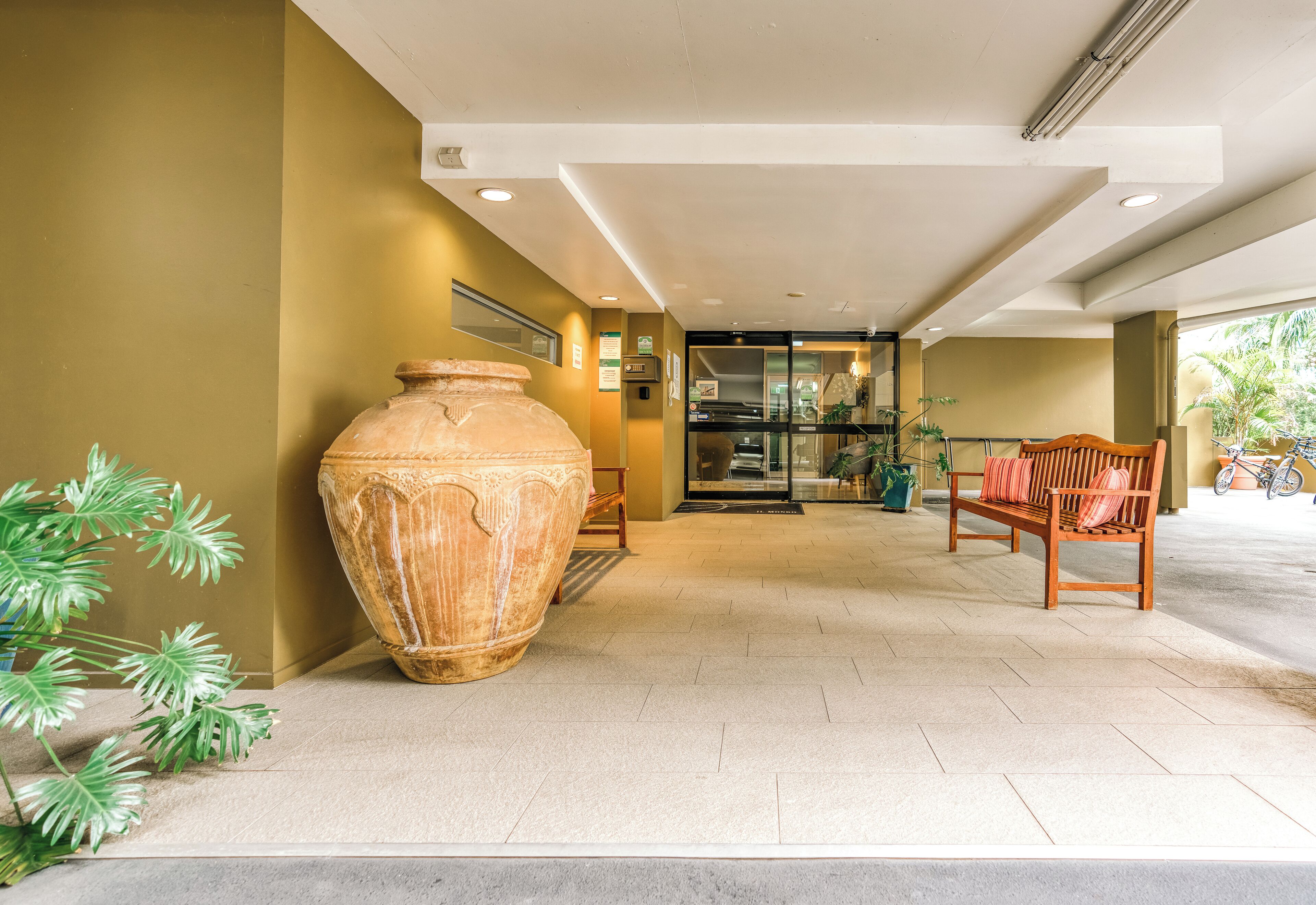 A spacious lobby with a large terracotta pot, a wooden bench, and a glass door leading to a room.