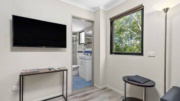 Una habitación de hotel moderna con un televisor de pantalla plana, una pequeña mesa con revistas, una mesita redonda con un libro, una ventana con vistas a árboles y una puerta que da a un baño.