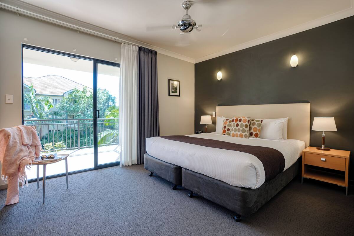 Une chambre d’hôtel avec un grand lit, une petite table avec une chaise, un balcon donnant sur un espace verdoyant et un ventilateur de plafond.