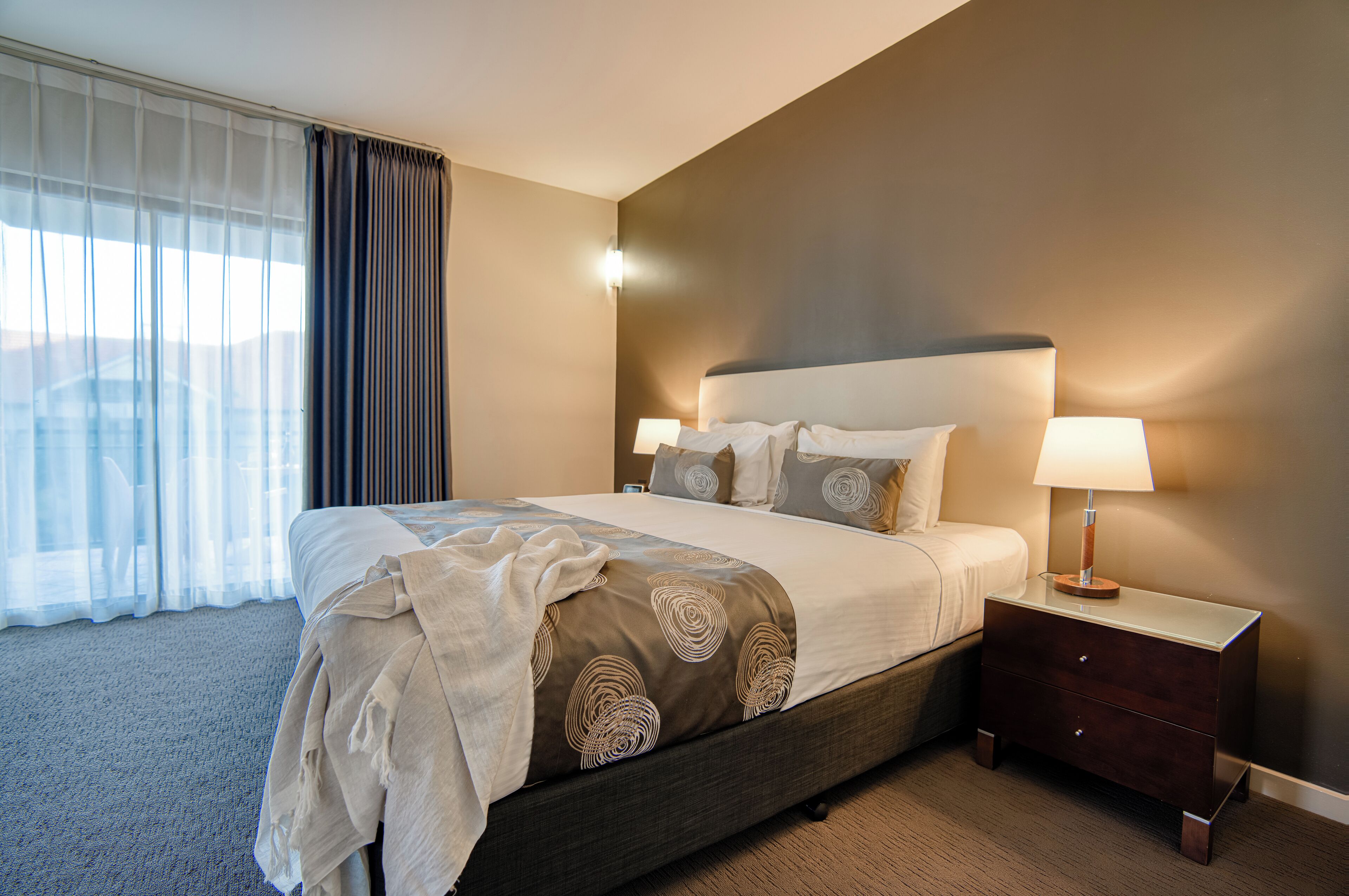Habitación de hotel con una cama grande, una mesita de noche con lámpara y vista a la ciudad por la ventana.