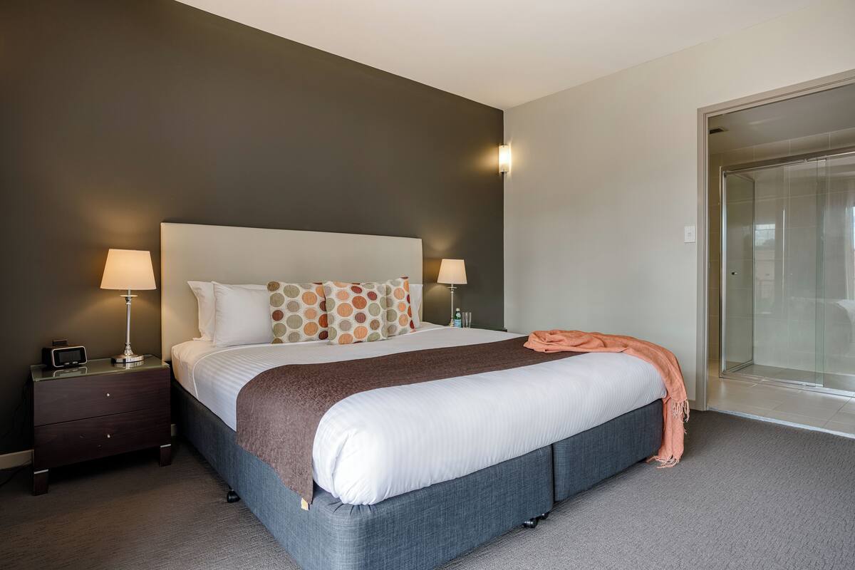 Une chambre d’hôtel moderne avec un grand lit, deux tables de chevet avec des lampes, une porte vitrée donnant sur un balcon et un mur de couleur sombre.