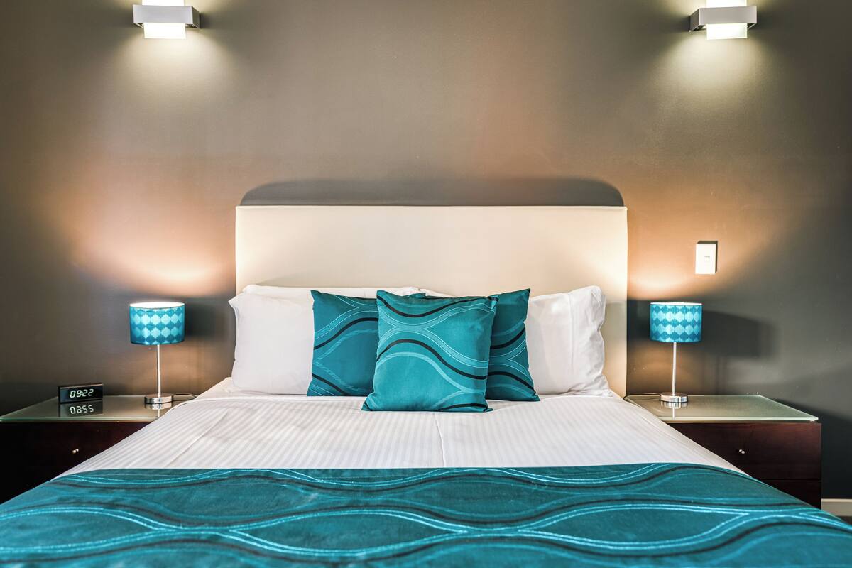 Un lit bien fait, recouvert d’une couvre-lit à motifs turquoise et agrémenté d’oreillers. Deux lampes de chevet aux abat-jour à motifs bleus, et une horloge numérique affichant l’heure.