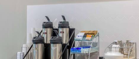 Una estación de café con dos máquinas de espresso, un dispensador de café y un espumador de leche. Hay vasos desechables y un molinillo de café en la encimera.