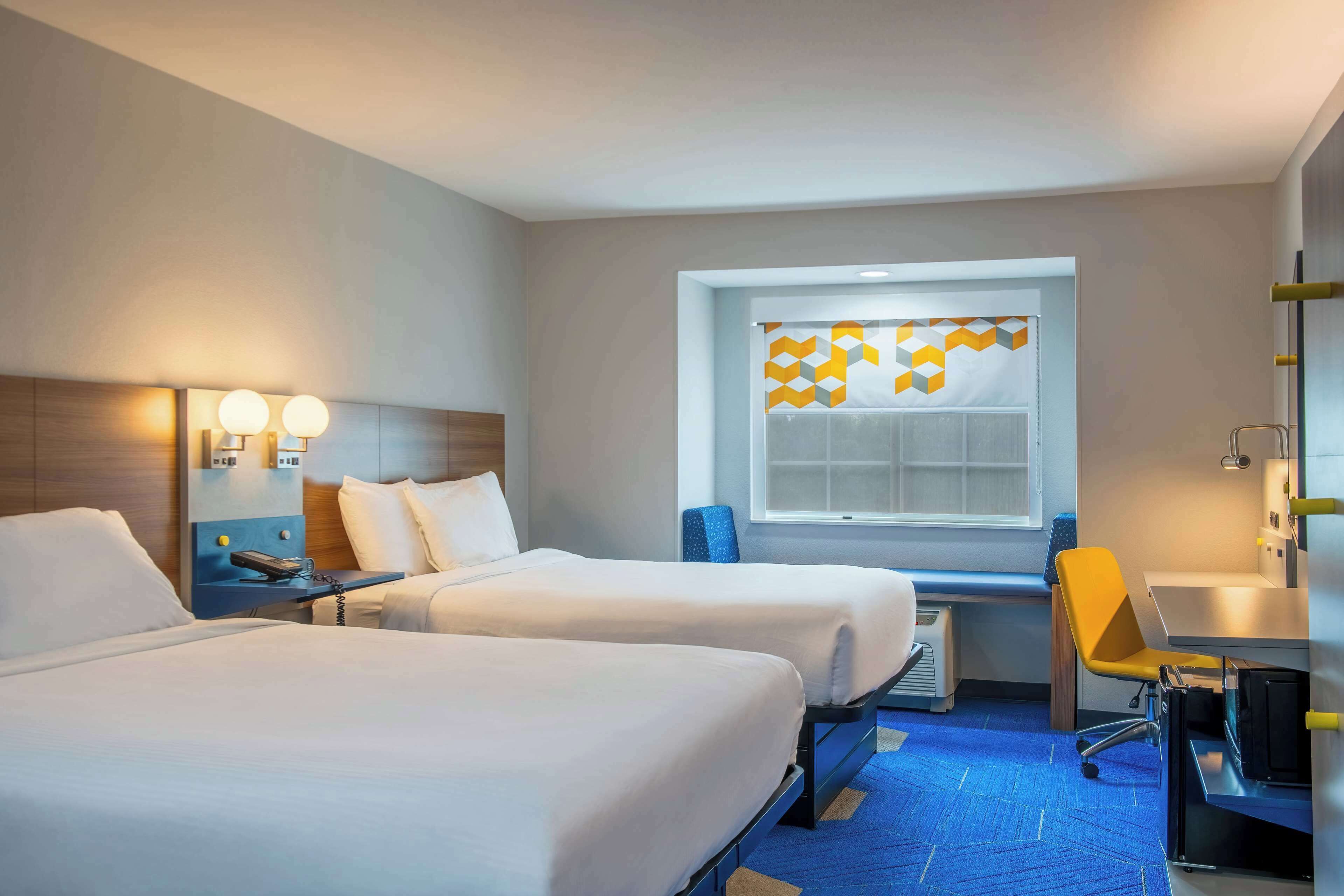 Habitación de hotel con dos camas, un escritorio, una silla, una ventana con cortina de diseño y una lámpara de pared.