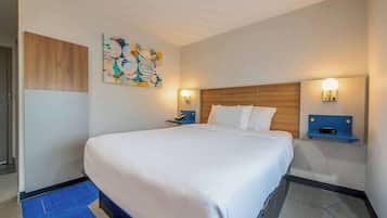 Une chambre d’hôtel avec un grand lit, deux tables de chevet avec des lampes, une tête de lit en bois, une peinture abstraite aux couleurs vives et un tapis bleu.