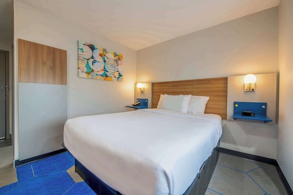 Une chambre d’hôtel avec un grand lit, deux tables de chevet avec des lampes, une tête de lit en bois, une peinture abstraite aux couleurs vives et un tapis bleu.