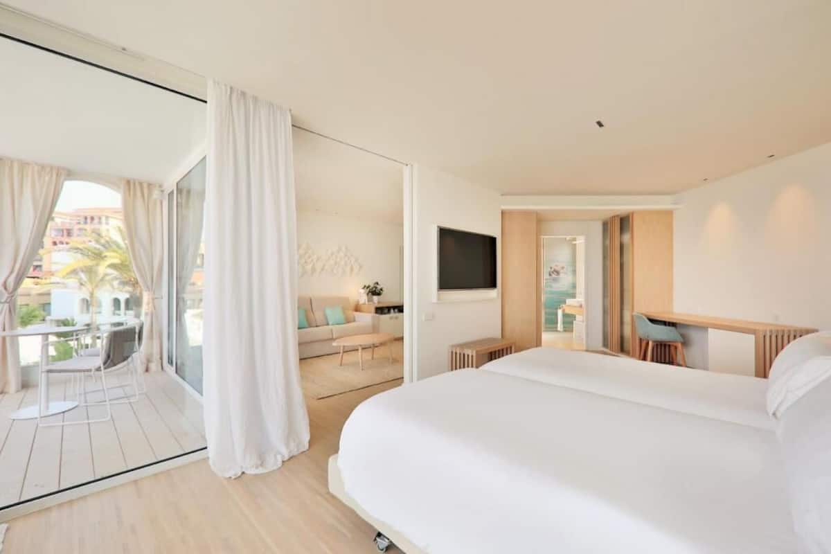 A modern hotel room with a large bed, a TV, a balcony with a table and chairs, and a view of the city.