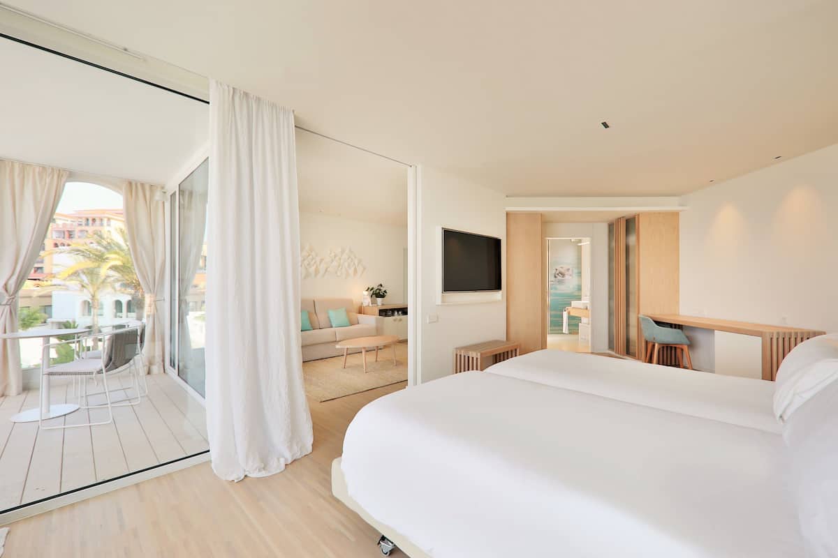A modern hotel room with a large bed, a TV, a balcony with a dining area, and a view of the city.