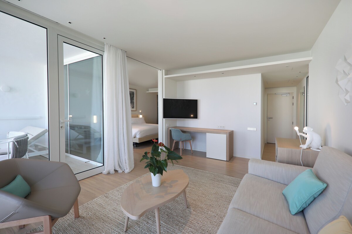 A modern hotel room with a glass door leading to a balcony, a flat-screen TV mounted on the wall, a desk with a chair, and a sofa with a cushion.