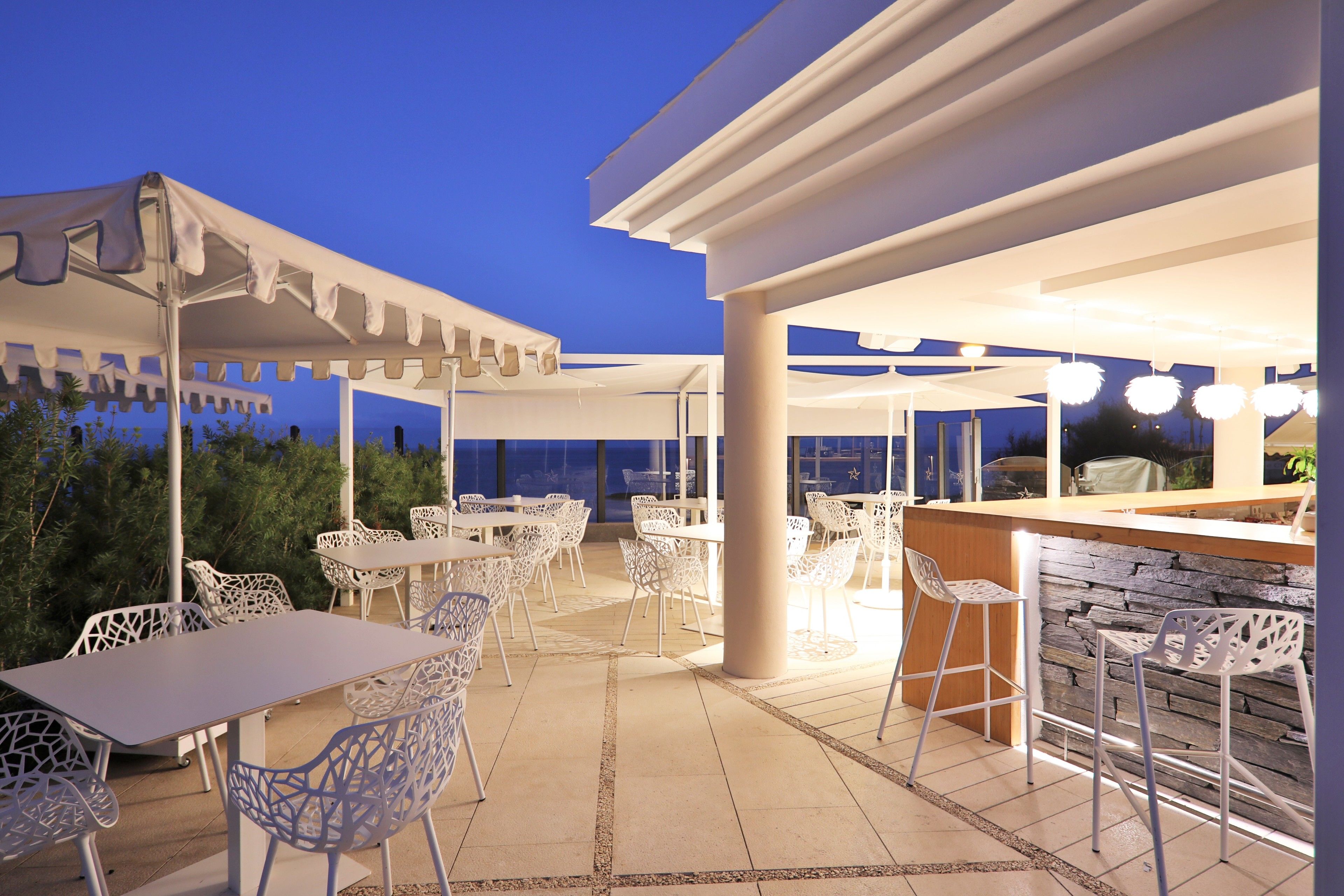 Un espace extérieur aménagé en terrasse, avec des chaises et des tables en métal blanc, un coin bar et une vue sur le ciel nocturne.