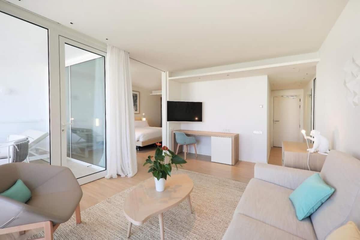 A modern hotel room with a large sliding glass door, a flat-screen TV, a desk, a sofa, and a small table with a plant.
