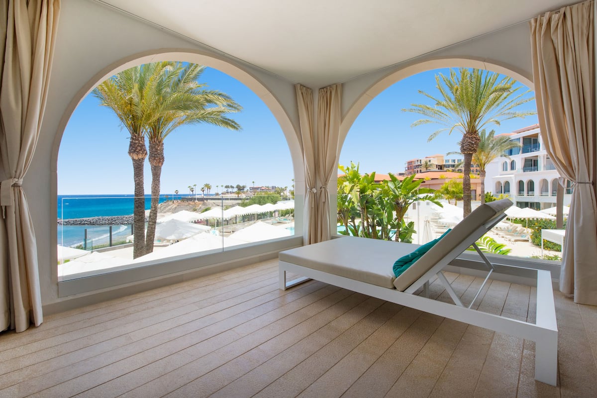 A room with a view of the sea, a lounge chair, and a large arched window.