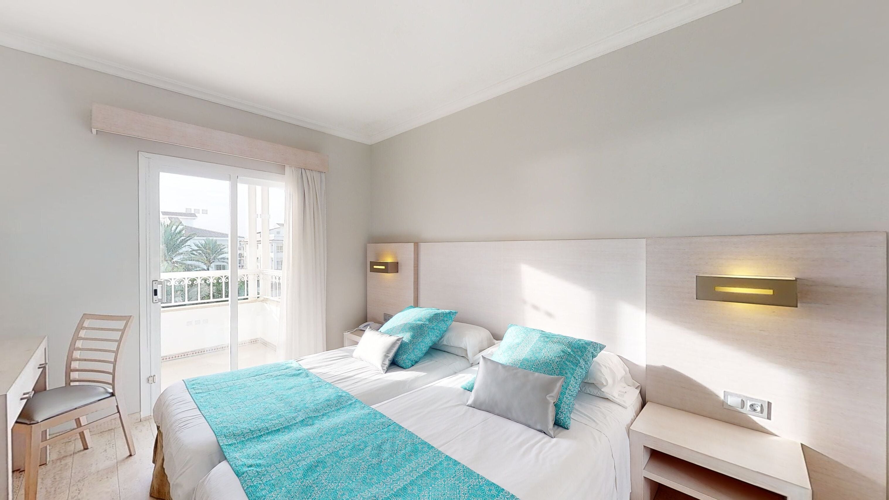 Ein Hotelzimmer mit einem großen Bett, zwei Nachttischen, einem Sessel und einem Balkon mit Blick auf Gebäude und Bäume.