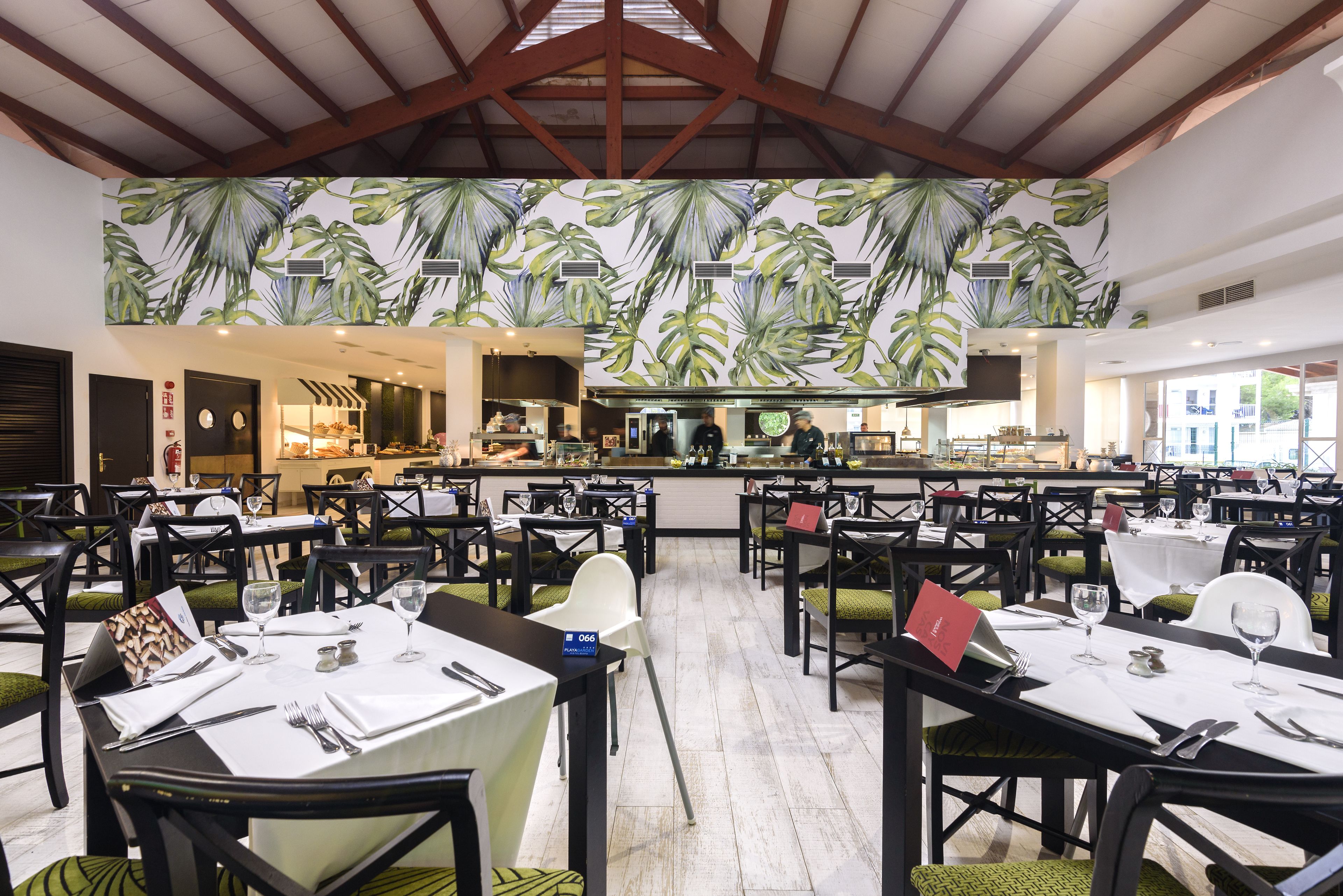Un restaurante con varias mesas dispuestas para comer, una zona de cocina al fondo y un diseño de hojas tropicales en el techo.