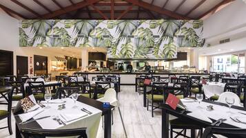 Un restaurante con varias mesas dispuestas para comer, una zona de cocina al fondo y un diseño de hojas tropicales en el techo.