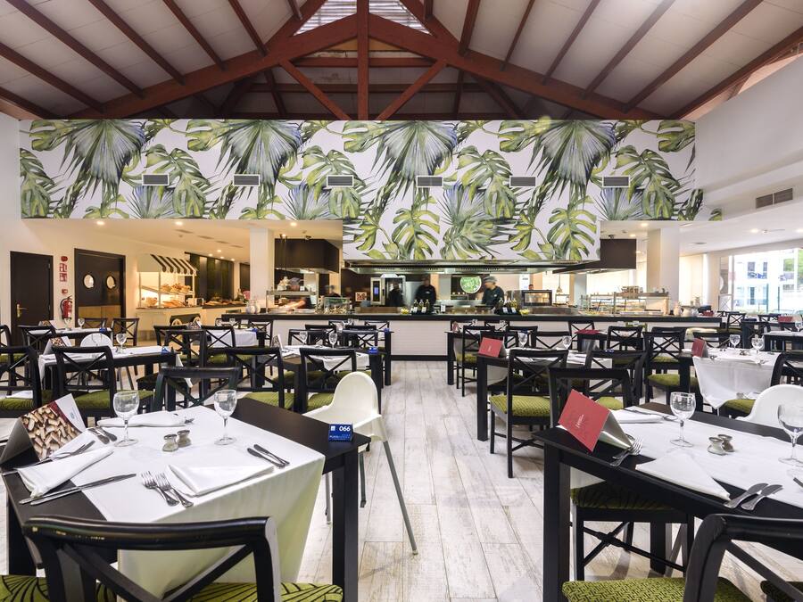 Un restaurante con varias mesas dispuestas para comer, una zona de cocina al fondo y un diseño de hojas tropicales en el techo.