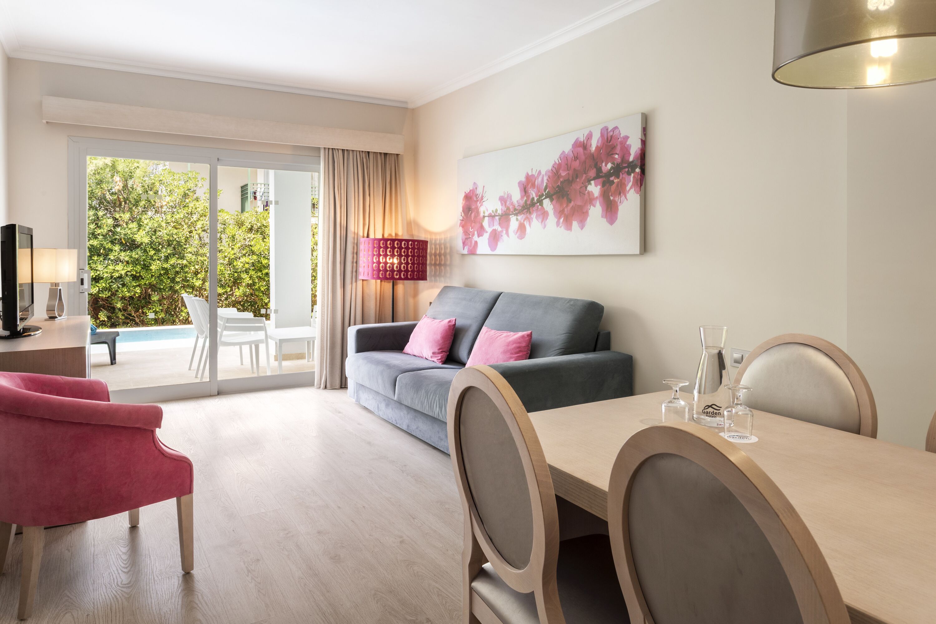 A modern living room with a grey sofa, pink cushions, a wooden dining table, and a large window with a view of greenery.